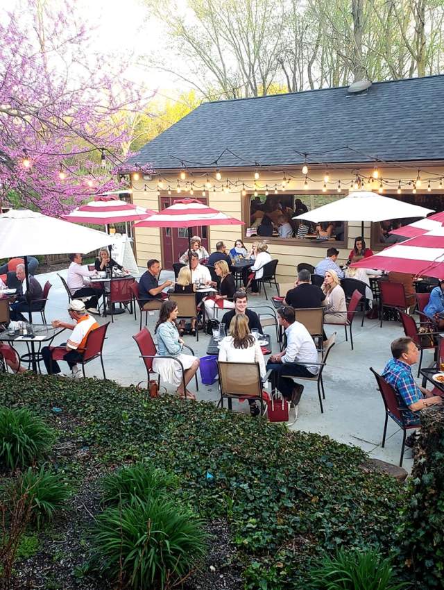 Outdoor patio at The Italian House on Park in Westfield, with diners enjoying food and drinks under red and white umbrellas surrounded by spring blooms and landscaping.