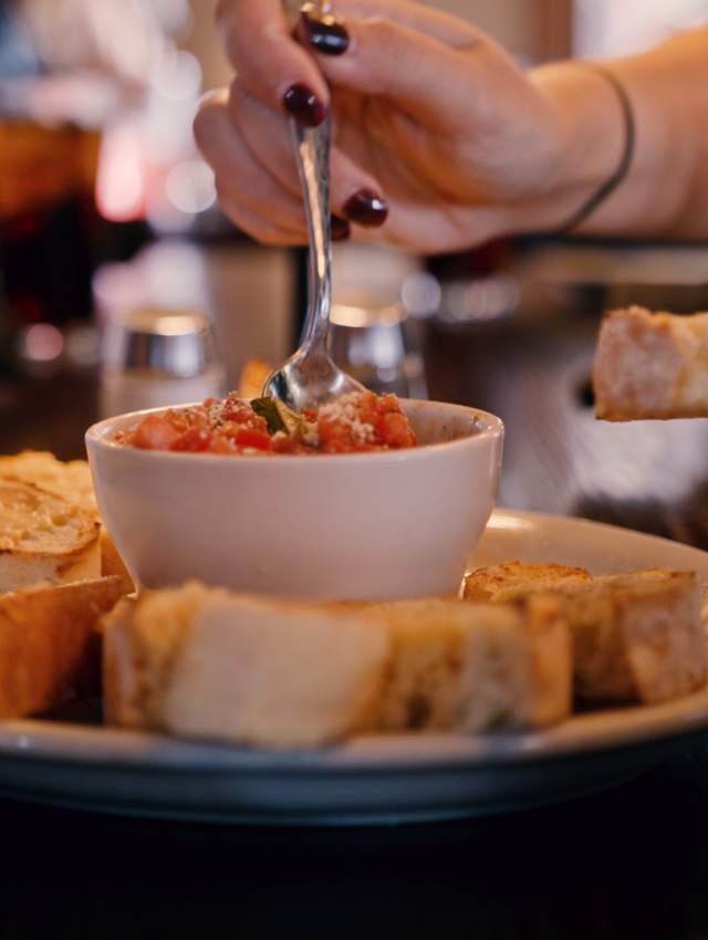 A plate of toasted bread slices surrounds a small bowl of tomato dip. A hand holds a spoon, ready to serve. Glasses of water are in the background.