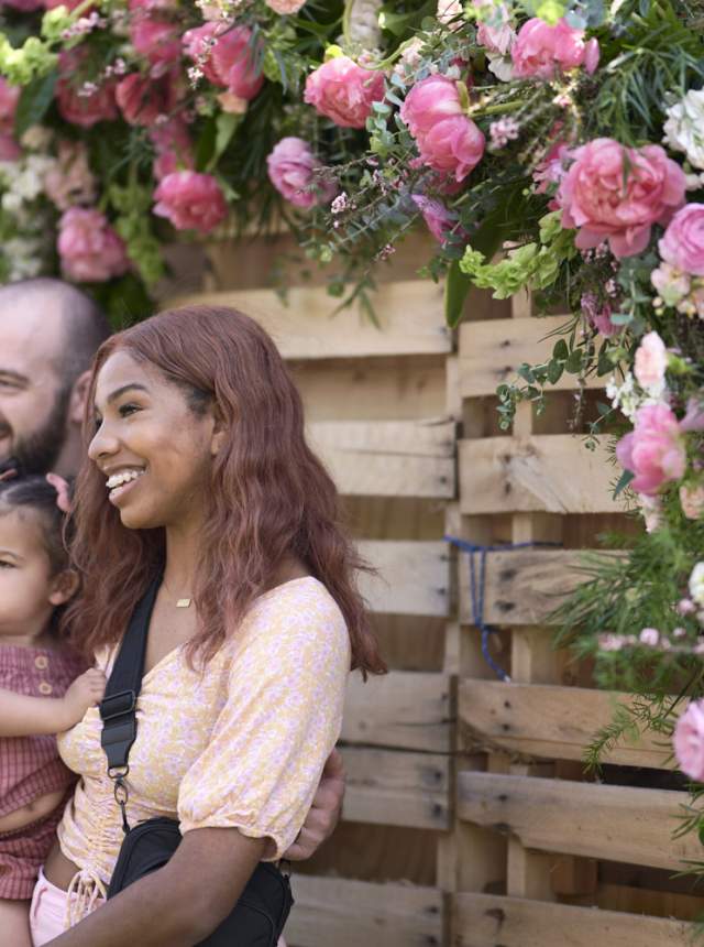 A smiling family of four poses in front of a wooden backdrop decorated with lush pink peonies at the Indiana Peony Festival. The parents hold their two young children as they stand beside the flower-covered wall.