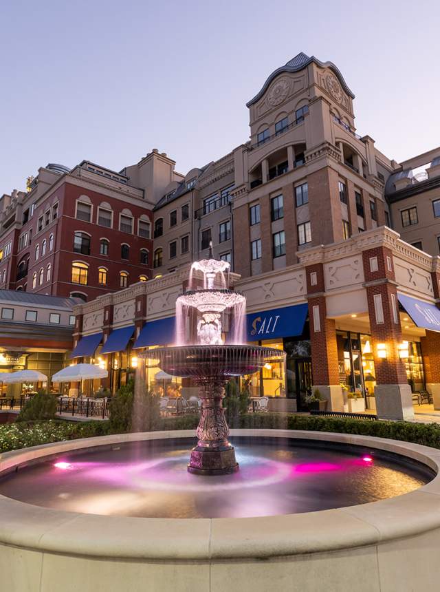 A mixed-use urban complex at dusk with multi-story brick and stone buildings lit from within. In the foreground, a small fountain with water flowing into a basin sits in a landscaped plaza, while storefronts and sidewalks line the street.