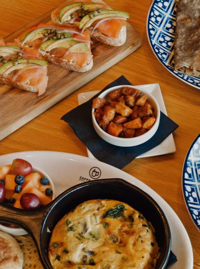 Brunch dishes arranged on a wooden table, including smoked salmon toast, a baked egg crêpe, waffles topped with powdered sugar, an omelet, fruit, potatoes, and a cup of coffee.