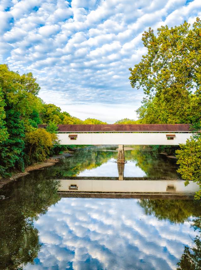 A covered bridge spans a calm river surrounded by lush green trees.