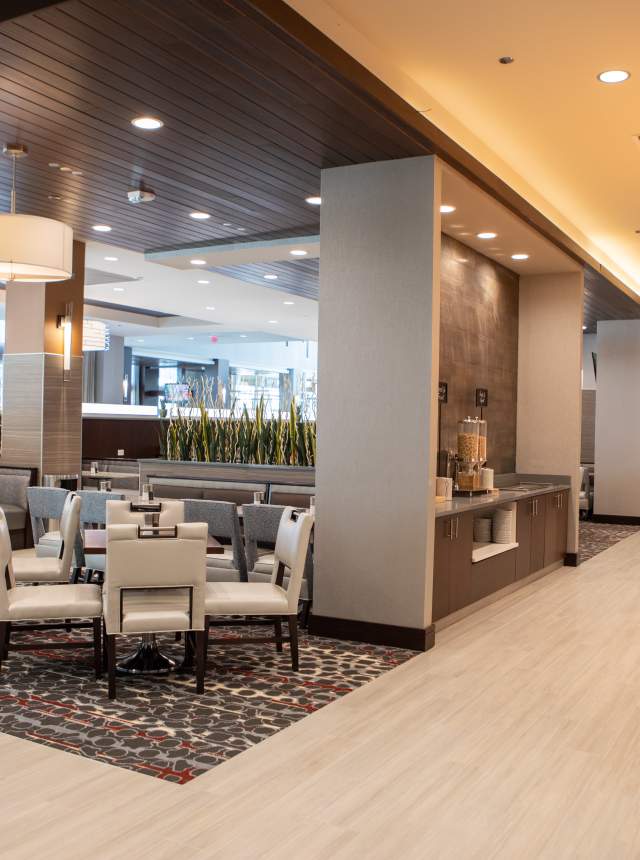 Modern hotel lobby featuring contemporary dining area with cream-colored chairs and patterned area rug in foreground, gray booth seating along windows, dark wood-paneled ceiling accents, white structural columns, and breakfast buffet service area with staff member visible in background at Embassy Suites Noblesville, Indiana.