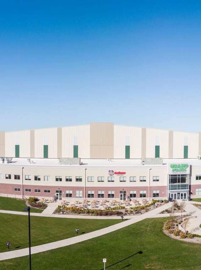 Wide aerial view of the exterior of the Grand Park Events Center, a large modern sports facility with a tan-and-white facade, brick lower level, green lawn, walking paths, and a central glass entrance under a clear blue sky.