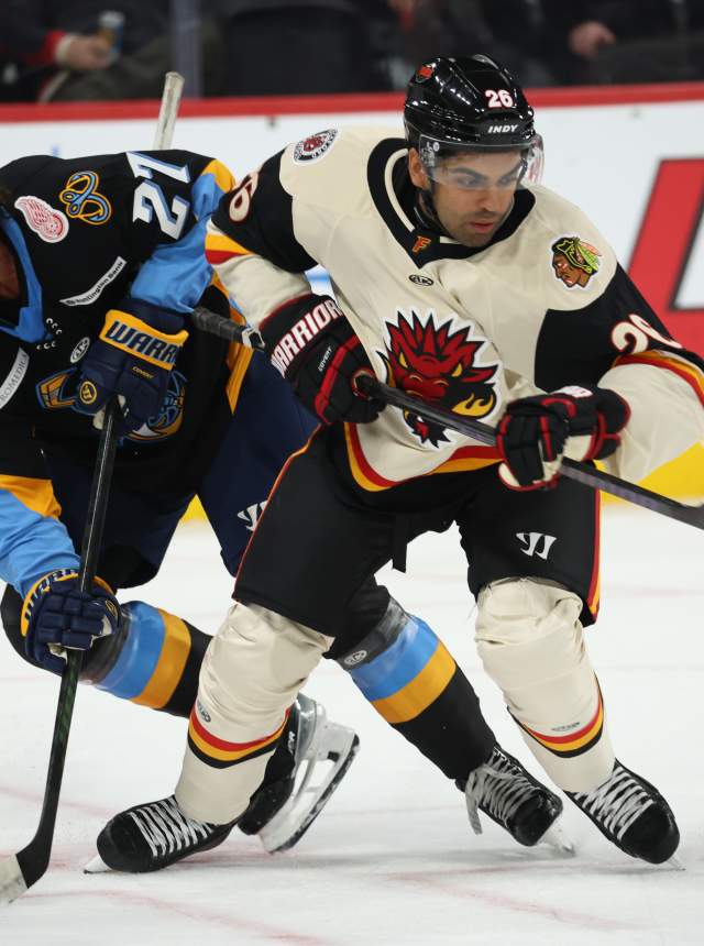Two ice hockey players compete for the puck on the rink, with a referee in the background. The arena features promotional signage.