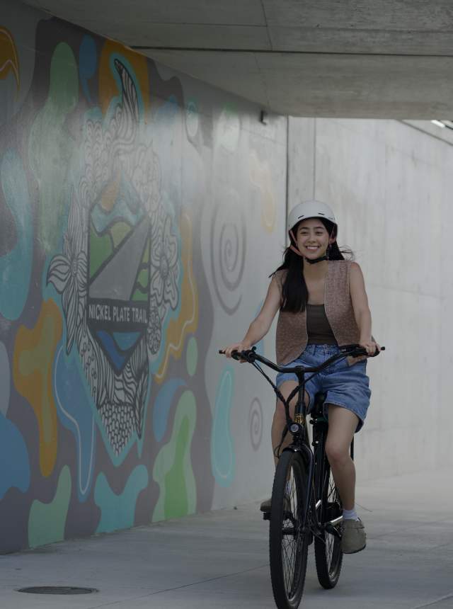 A woman wearing a helmet smiles while riding a bicycle through an underpass featuring a colorful Nickel Plate Trail mural on the wall beside her.