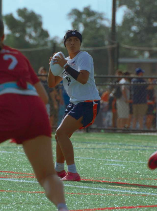 Outdoor flag football game on a turf field with multiple players in motion, including one player in the center preparing to throw the ball while others run routes and defenders move in to cover, with spectators and fencing visible in the background.