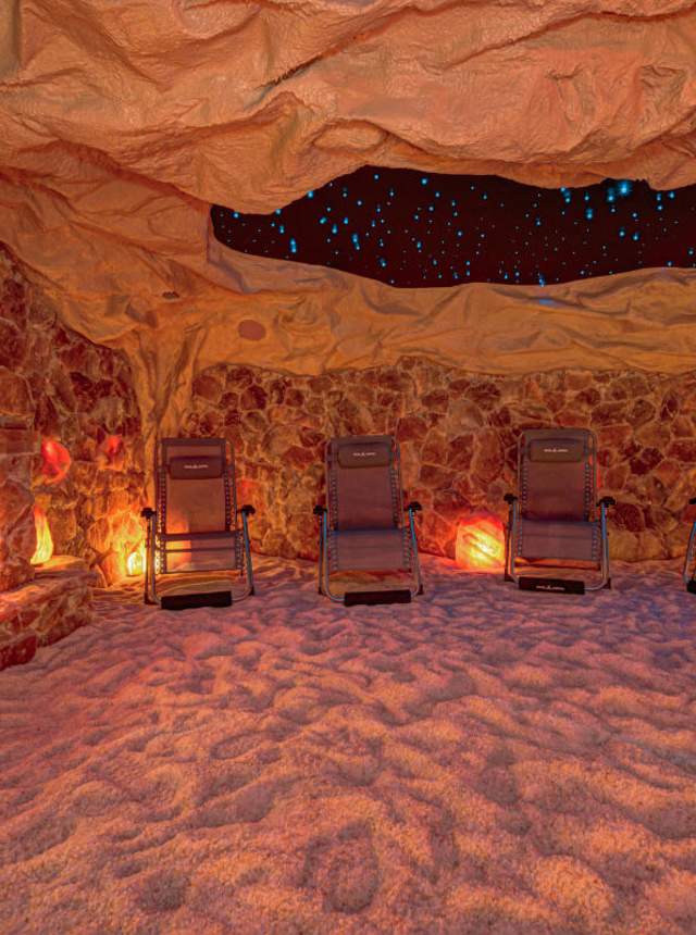 A room designed to look like a cave, with stone-textured walls, a sandy floor, glowing salt lamps, a faux fireplace on the left, and several reclining lounge chairs arranged in a semicircle.