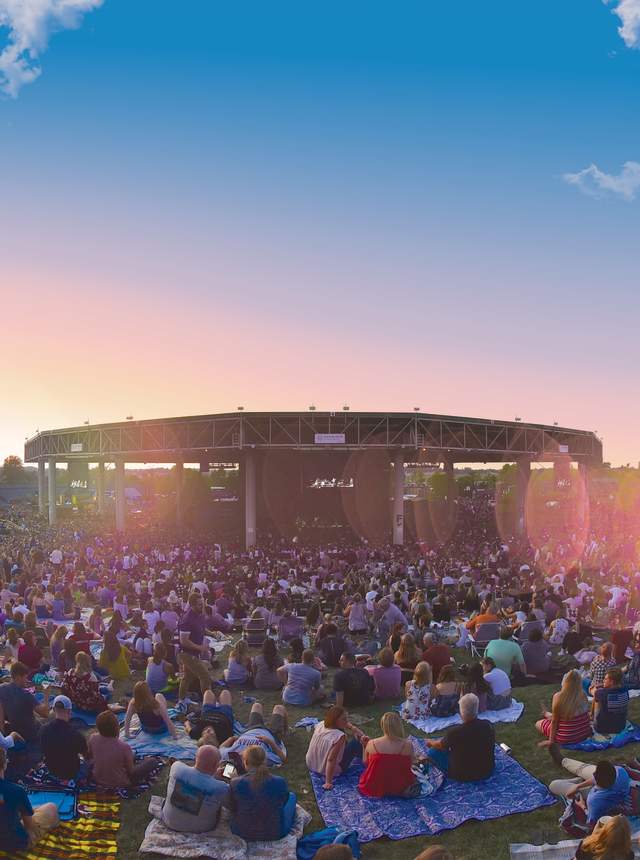 A large crowd gathers on the lawn at Ruoff Music Center at sunset, facing the outdoor stage as colorful sky and lens flare frame the scene.