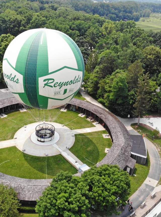 Hot air balloon in vibrant green and white colors floating above a grassy field, trees and a barn below