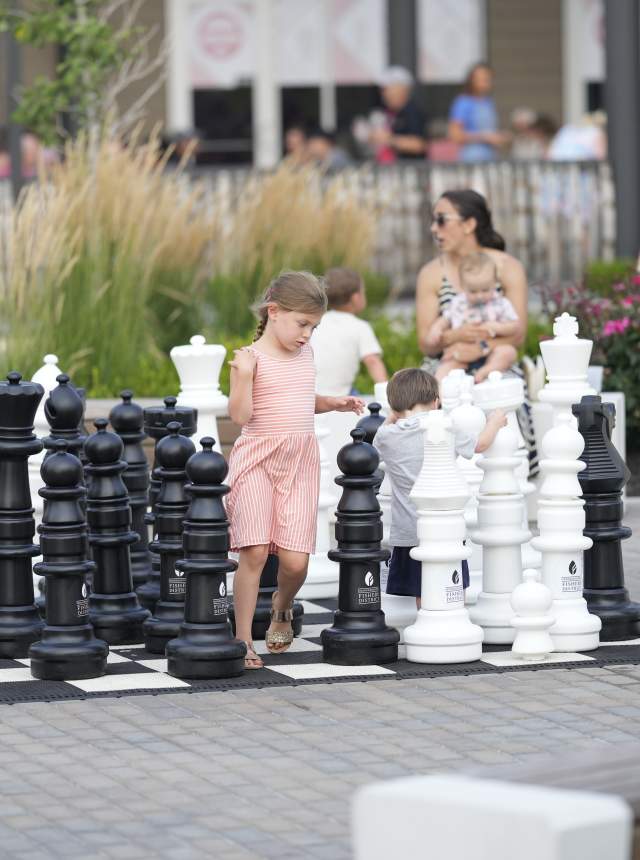 Children and adults stand around a large outdoor chess set with oversized black and white pieces arranged on a paved plaza, with plants and buildings in the background.