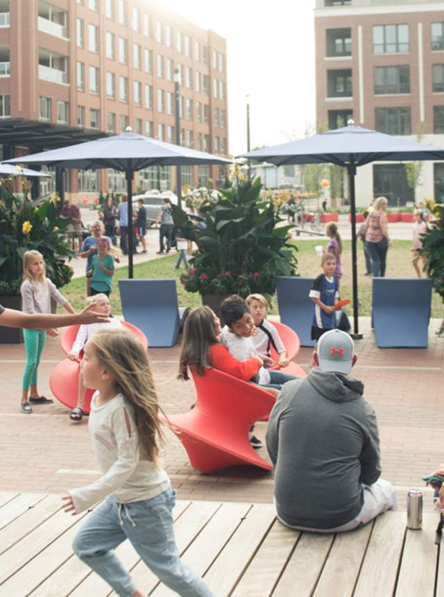 A lively urban street with people walking, children playing, outdoor dining, and brick buildings
