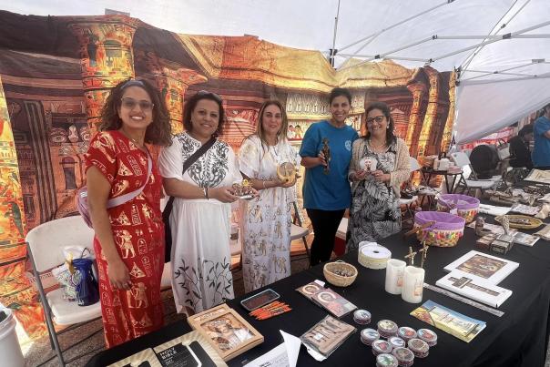 Five people stand behind a vendor table displaying crafts and artwork at the Egyptian Festival, with a backdrop depicting ancient Egyptian architecture.