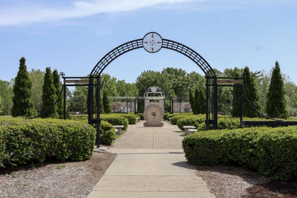 A decorative archway leads into a landscaped garden with neatly trimmed hedges and a central stone feature, under a clear blue sky.