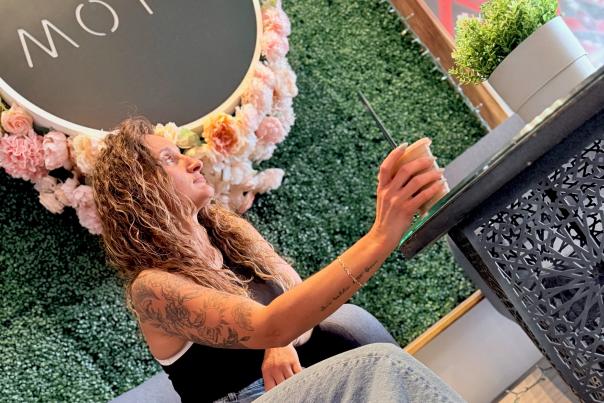 Deaf blogger Irina Burgess sitting in MOTW coffee shop in front of a sign on a greenery wall backdrop.