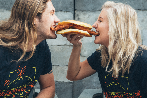 Two people smiling while sharing a breaded pork tenderloin sandwich, leaning toward each other.