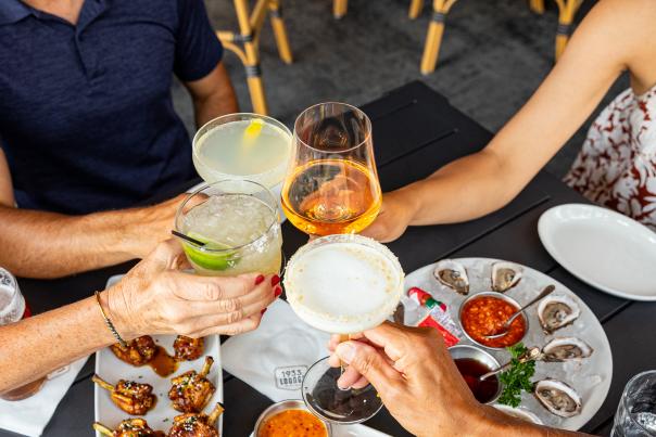 Overhead view of a group at an outdoor restaurant table raising cocktails, wine, and a pint of beer in a toast, surrounded by plates of oysters on ice, fried calamari, grilled lamb chops, and dipping sauces.