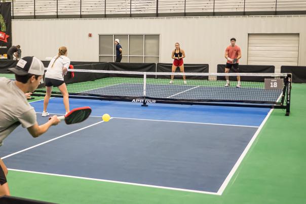 A player serves a yellow ball on a blue pickleball court, with spectators and officials observing in a spacious indoor facility.