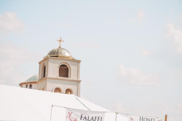 Outdoor scene at the St. George Greek Festival, where guests visit food tents labeled ‘Falafel’ and ‘Honey Balls.’ The church’s domed tower rises in the background under a clear sky.