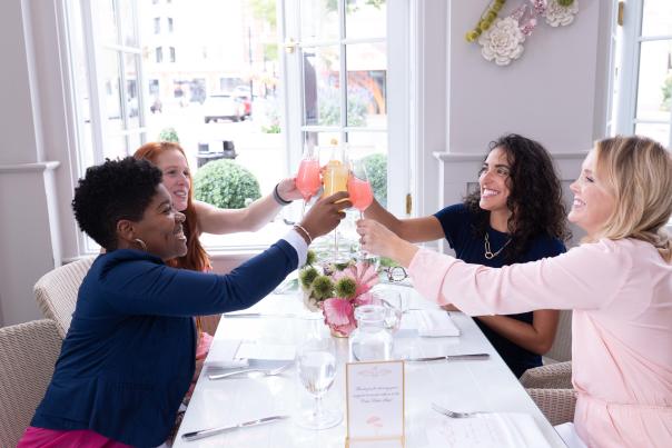 Four people sit around a white table in a bright dining room, raising glasses in a toast, with place settings, flowers, and large windows in the background.