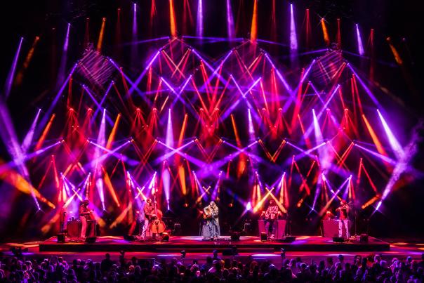 Band performing on a large stage with vibrant purple, red, and orange light beams crisscrossing above them, viewed from a packed audience.