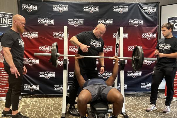 A football scouting combine participant performs a bench press while lying on a weight bench, surrounded by three staff members spotting and observing in front of a branded combine backdrop.