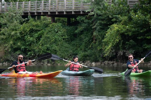 Three people wearing life jackets paddle kayaks along the White River, with trees and a wooden bridge in the background.
