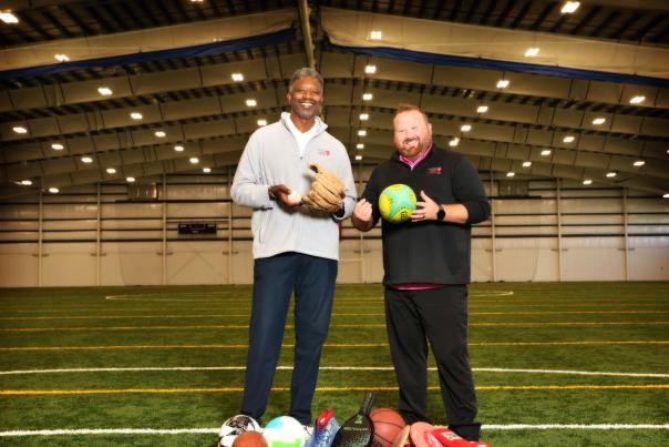 Two men standing on an indoor turf field holding sports balls, with additional balls on the ground in front of them. Rows of lights line the ceiling of the large sports facility behind them.