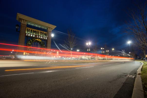 Illuminated Hamilton Town Center entrance at night with holiday decor, as long-exposure car light trails streak along the curved road under a deep blue sky.