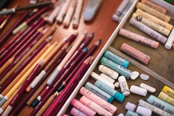 A close-up view of assorted artist tools arranged on a wooden surface, including colored pencils, paintbrushes, and pastel sticks sorted into trays by color and type.