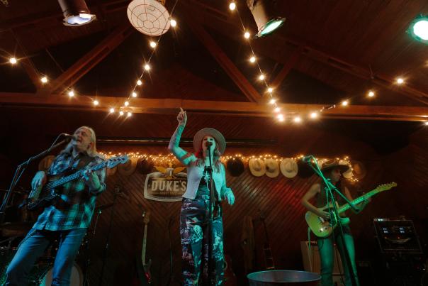 A band performs on a dimly lit indoor stage decorated with string lights. The lead singer stands at the microphone with one arm raised, while two guitarists play on either side.