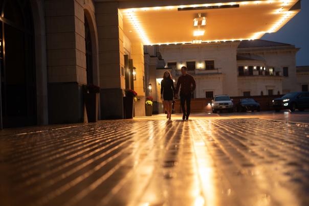 A couple walks hand-in-hand on a wet, reflective pathway under warm lights outside a grand hotel entrance at dusk.