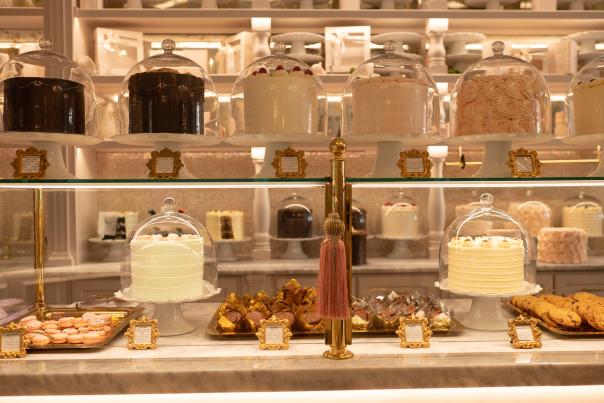 A bakery display case at Cake Bake showcases an elegant arrangement of desserts and ingredients. Glass jars filled with baking staples line the back shelves, while the front counter features neatly arranged pastries, cookies, and small cakes displayed on trays and under glass domes. Warm lighting, gold accents, and symmetrical styling create a refined, upscale atmosphere that highlights the bakery’s decorative presentation and artisanal treats.