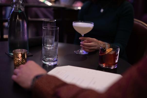 Dimly lit lounge table with two people seated; one holds a pale cocktail in a coupe glass, while an old fashioned, water glasses, and a candle sit on the table.