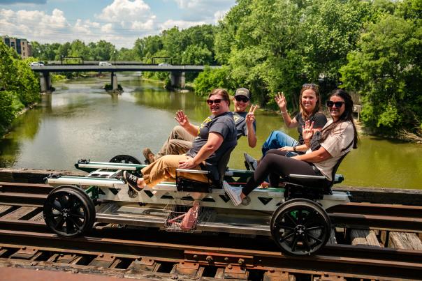 Four people ride a rail bike across a bridge, smiling and waving as they travel over a river surrounded by lush green trees.