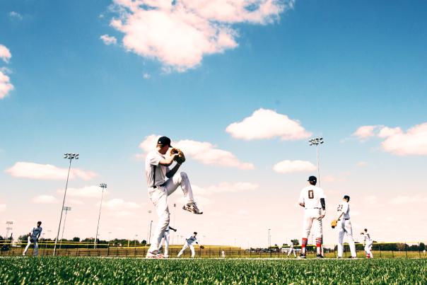 Baseball players in white uniforms on an outdoor field, with one player mid-throw in the foreground and others spread across the field under a bright blue sky with scattered clouds.