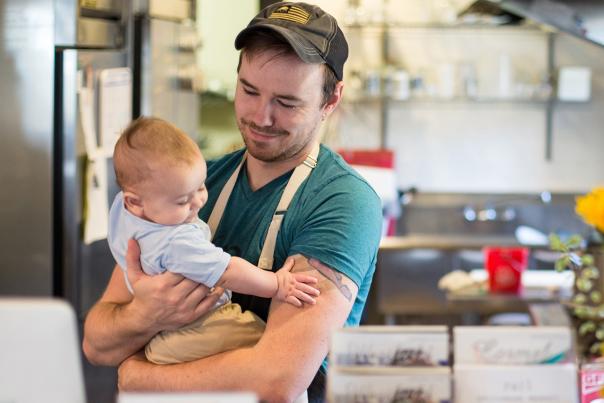 A man in an apron holding a baby in a professional kitchen setting.