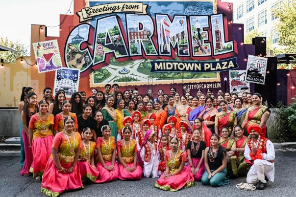 A large group of people in colorful traditional Indian attire pose together in front of the vibrant ‘Greetings from Carmel – Midtown Plaza’ mural during Diwali celebrations.