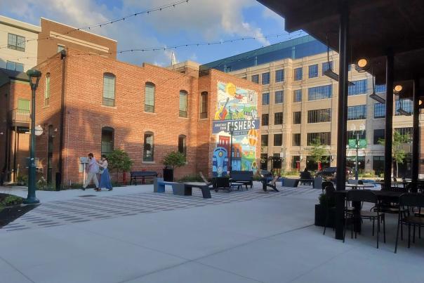 Open-air plaza in downtown Fishers, Indiana, featuring red-brick buildings, modern office buildings, and string lights overhead beneath a blue sky with large white clouds. A colorful “Greetings from Fishers” mural stands in the center near benches and outdoor seating, while a few pedestrians walk across the spacious paved courtyard.