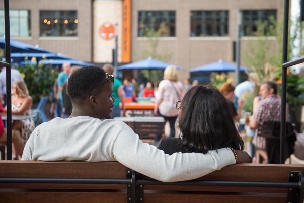 A couple sits close together on a bench at Midtown Plaza, the man’s arm around the woman as they look out over a lively crowd gathered around outdoor tables and blue umbrellas.
