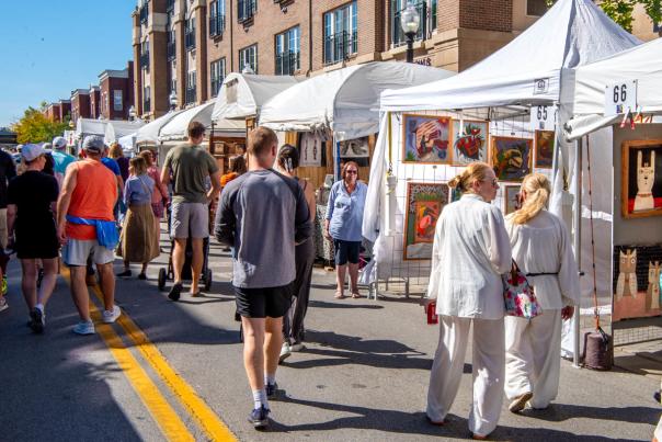 Crowds walk between rows of white artist booths at the Carmel International Arts Festival, browsing colorful artwork displayed along a sunny downtown street.