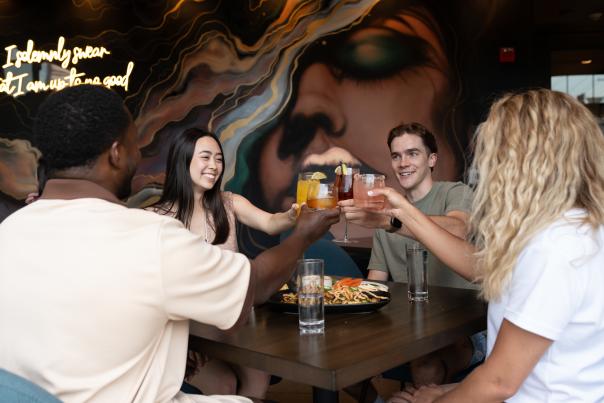 A group of four friends toasting with colorful drinks at a table, surrounded by a vibrant mural and plates of food.