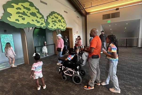 Visitors gather in a spacious indoor exhibit area with high wood-paneled ceilings and carpeted floors. A decorative entrance shaped like two large green trees frames an interactive display, while children and adults stand nearby, including a wheelchair user. A digital sign on the left reads “Your Journey Begins,” and several people are exploring the attraction.