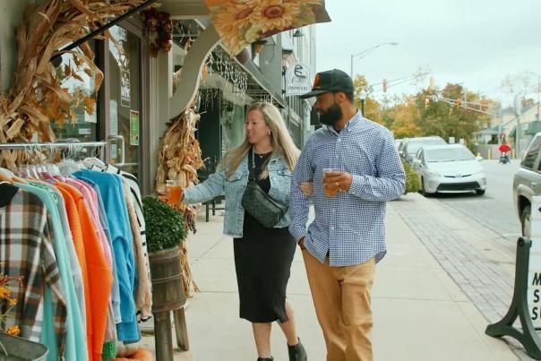 Two people stroll past a boutique storefront in downtown Noblesville, looking at a rack of colorful clothes on the sidewalk.