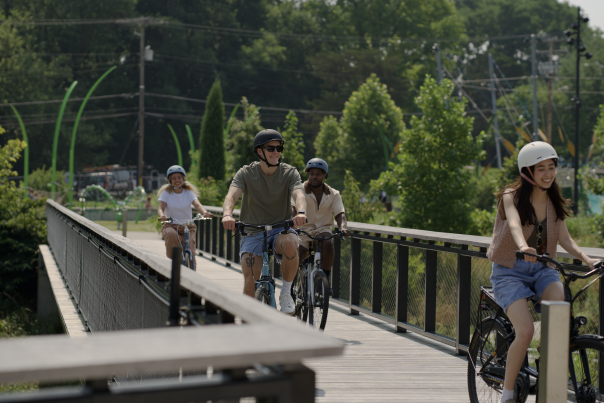 A group of four people wearing helmets ride bicycles across a wooden bridge at Grand Junction Plaza, surrounded by trees and greenery.