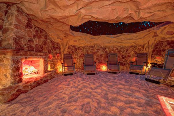 A room designed to look like a cave, with stone-textured walls, a sandy floor, glowing salt lamps, a faux fireplace on the left, and several reclining lounge chairs arranged in a semicircle.