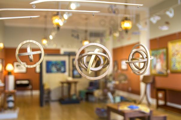 Hanging wooden sculptures in a bright art gallery, with colorful paintings on the walls and warm lighting creating an inviting atmosphere.