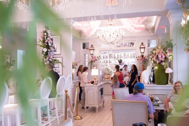 Brightly lit café interior with elegant chandeliers, floral decorations, and patrons engaging at the counter and tables. A welcoming atmosphere.