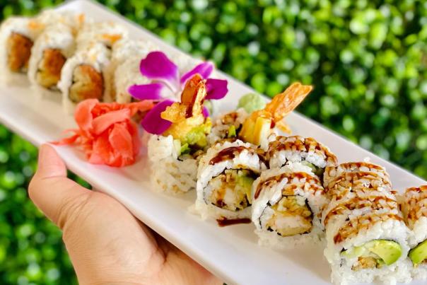 Assorted sushi rolls on a white platter with pickled ginger and floral garnish, held in front of a green foliage backdrop.