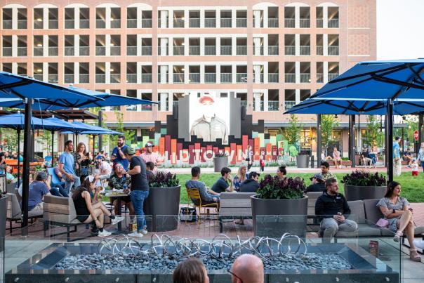 People relax and socialize around outdoor seating and fire features at Carmel’s Midtown Plaza, with large umbrellas, public art, and a tall office building in the background.